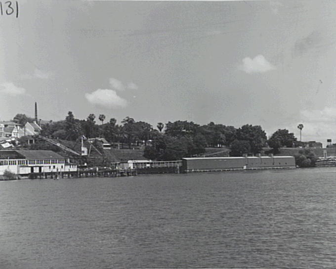 Black and white image from the water showing the pool and the box factory buildings on the cliff