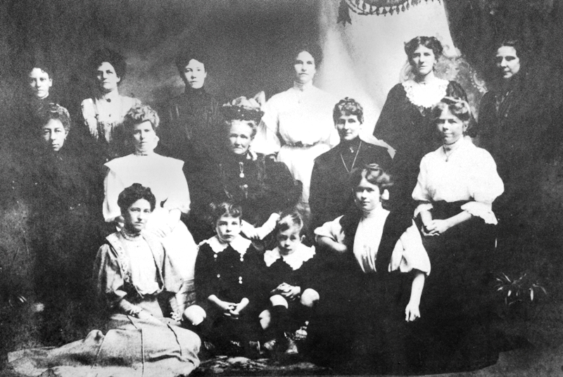 Black and white photograph showing a 13 people standing and sitting in Victorian style frilled dresses and hair. With two young boys crouched down in front