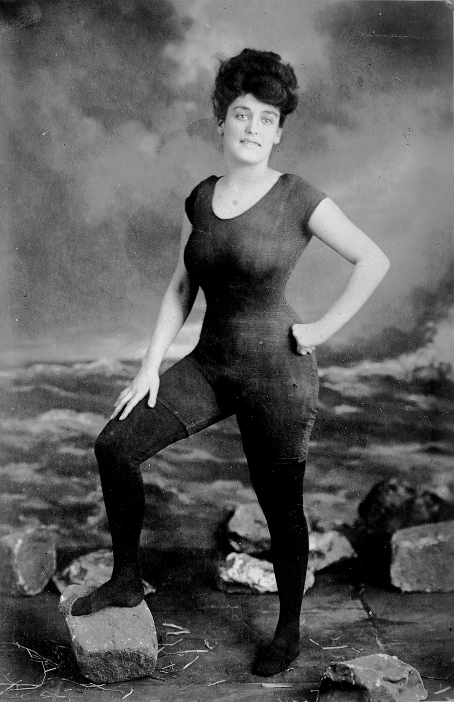 Black and white photograph of Annette Kellerman standing in a pose with left hand on hip in from of a fake photo studio to look like a rocky beach view. Wearing the controversial one piece swimsuit 