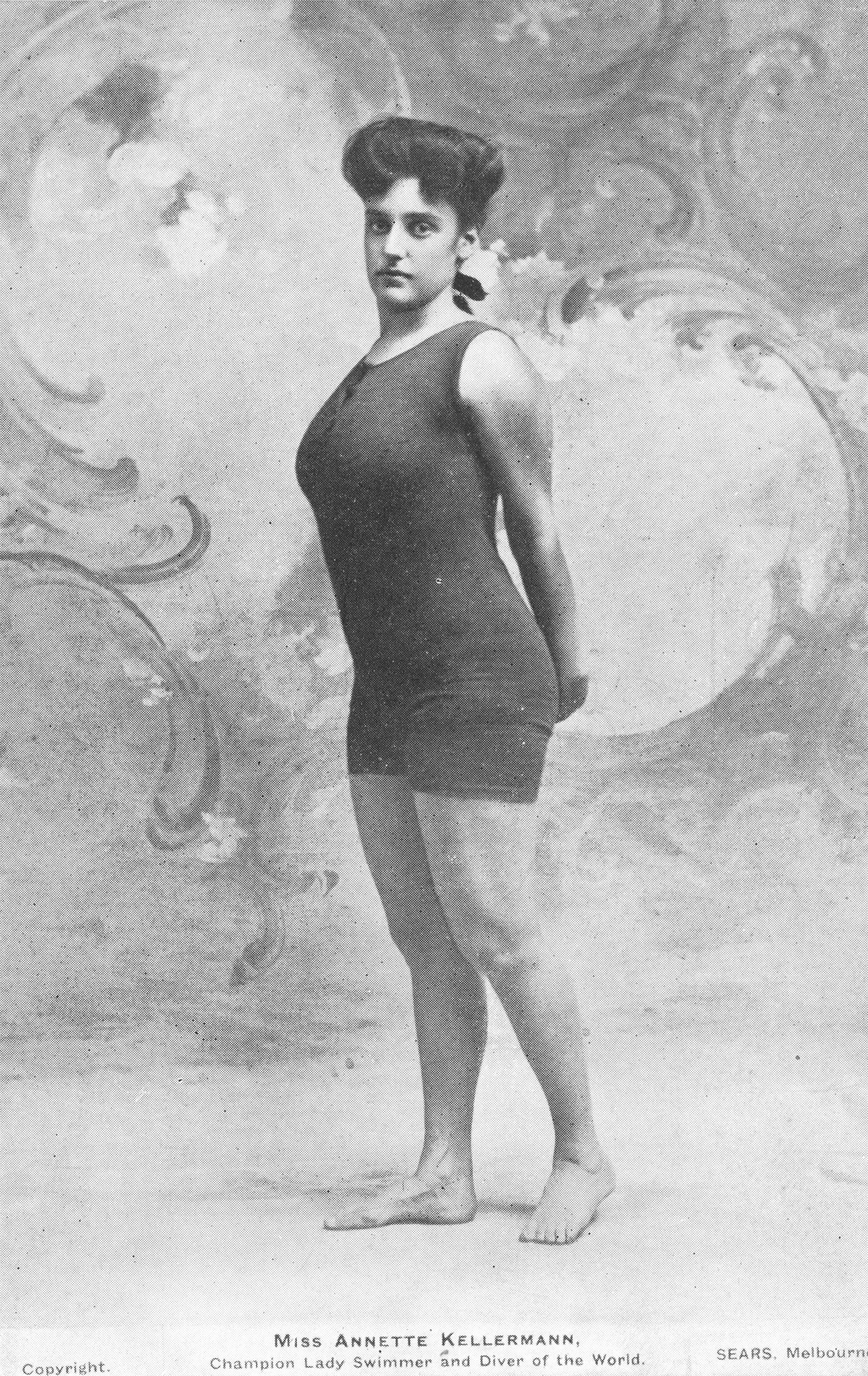 Black and white photograph of Annette Kellerman standing in a pose with hands behind back. Wearing the controversial one piece swimsuit.