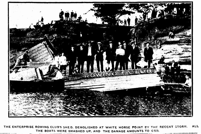 Black and white newspaper photo of the rowing club shed flatten from a storm, the front sign with name lays slightly off the base intothe water with 9 members posed behind it. a few row boats situated on the side. The roof appears in the midground collapsed with people all standing of the white horse point cliffside in background.